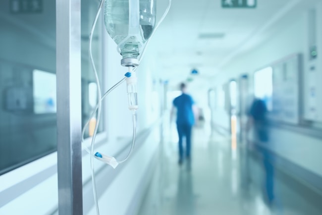 sjukhuskorridor med droppställning i förgrunden och blåklädd personal i bakgrunden