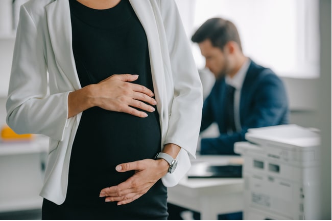 gravid kvinna på kontor med en man vid skrivbordet i bakgrunden