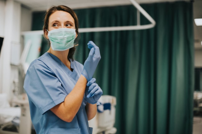 Kvinnlig undersköterska med munskydd sätter på sig blå latexhandskar.