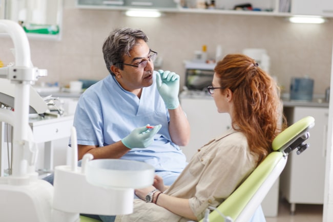 Manlig tandläkare pratar med kvinnlig patient som sitter i tandläkarstolen.