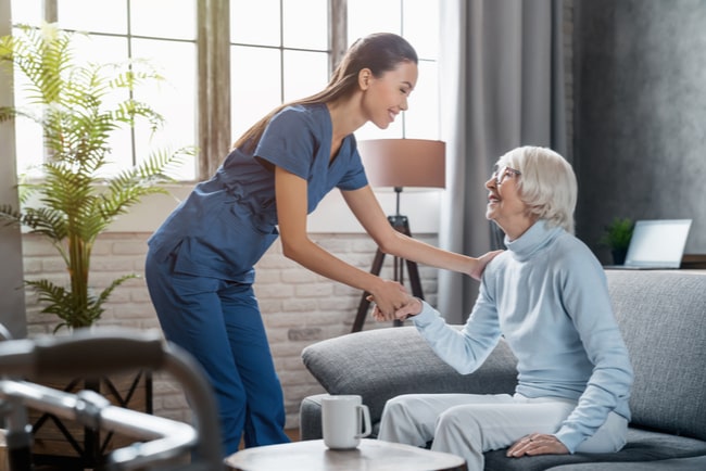 Ett vårdbiträde hjälper en äldre kvinna upp från en soffa.